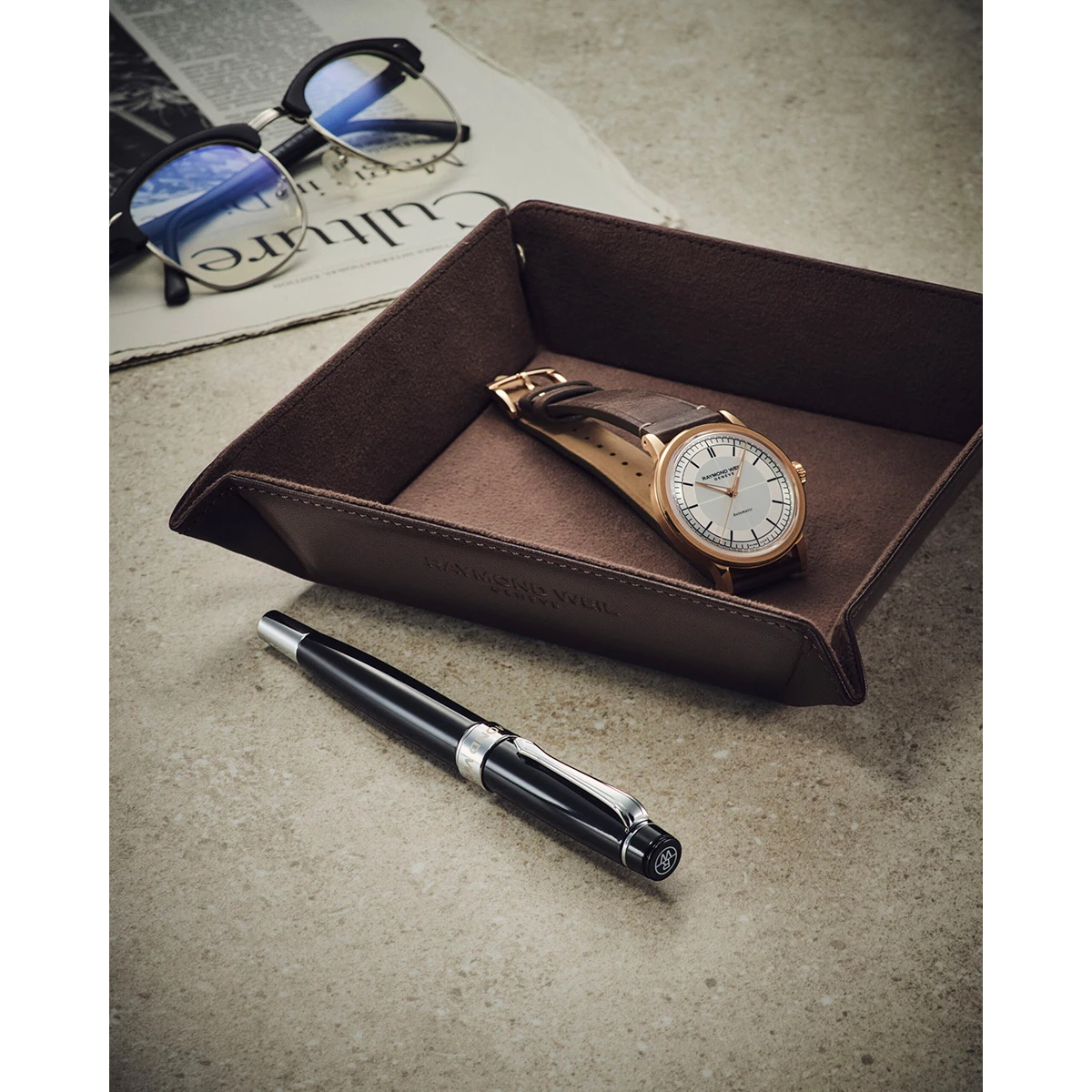 腕時計、ロゴ入りのレザー製レザートレイ、および万年筆が並んだイメージカット